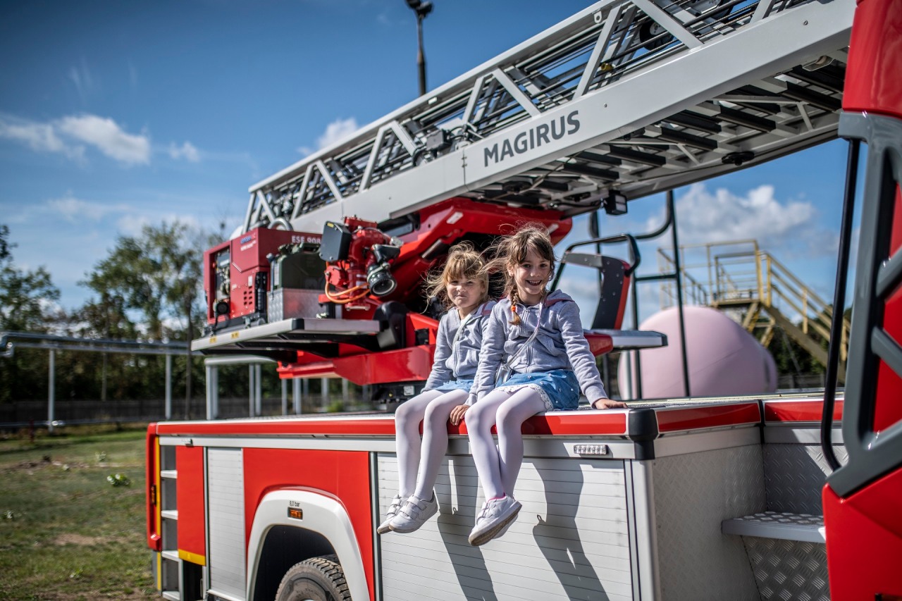 Fundacja OLREN. Dwie uśmiechnięte dziewczynki siedzą na karoserii wozu strażackiego. W tle teren rafinerii